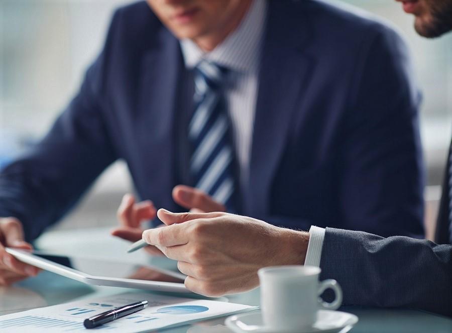 Due uomini in abiti formali discutono documenti e grafici a un tavolo con una tazza di caffè.