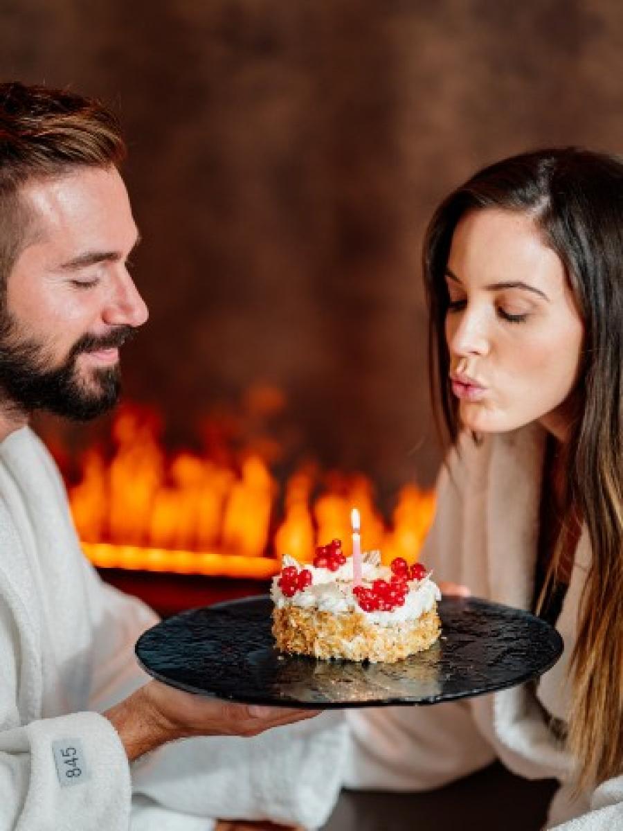 Donna che spegne una candela su una torta sorretta da un uomo, con un camino acceso sullo sfondo.