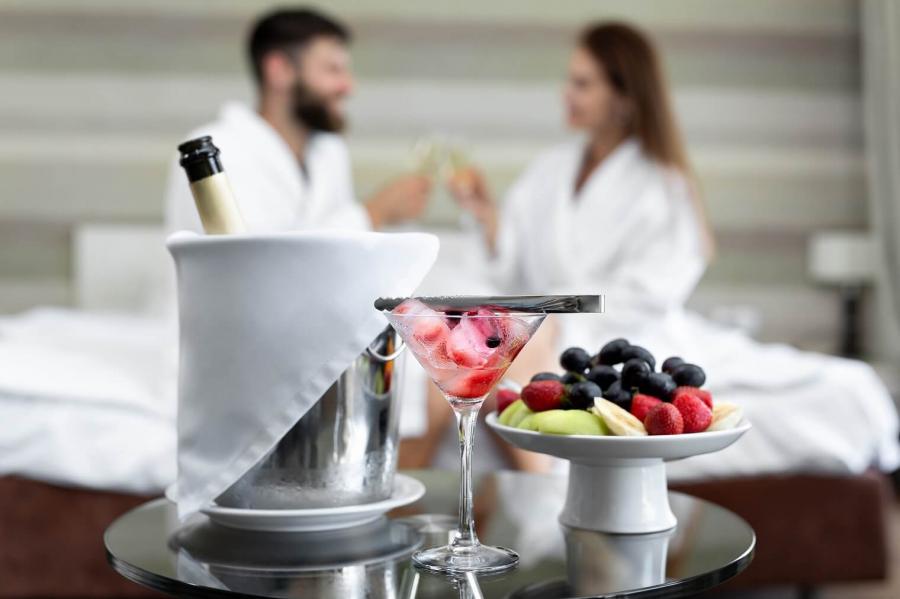 Coppia in accappatoio brinda con champagne davanti a frutta e cocktail in hotel.