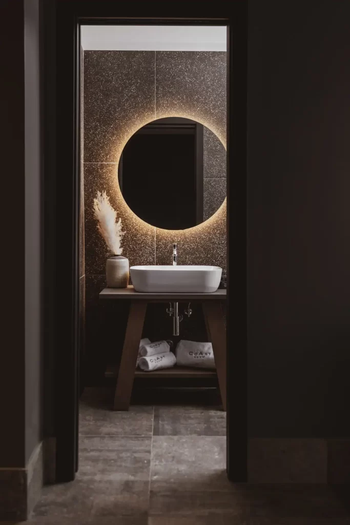 Bagno elegante con specchio circolare illuminato e lavandino bianco.