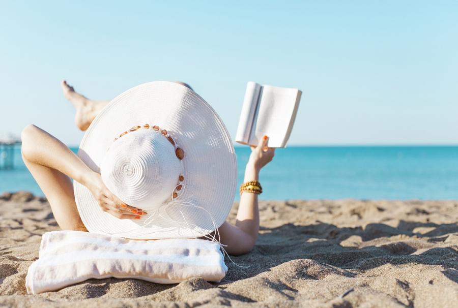 Donna con cappello bianco legge un libro sulla spiaggia al sole.