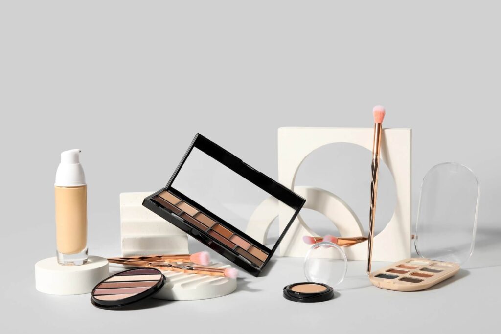 Prodotti cosmetici vari su fondo grigio, tra cui palette di ombretti e fondotinta.