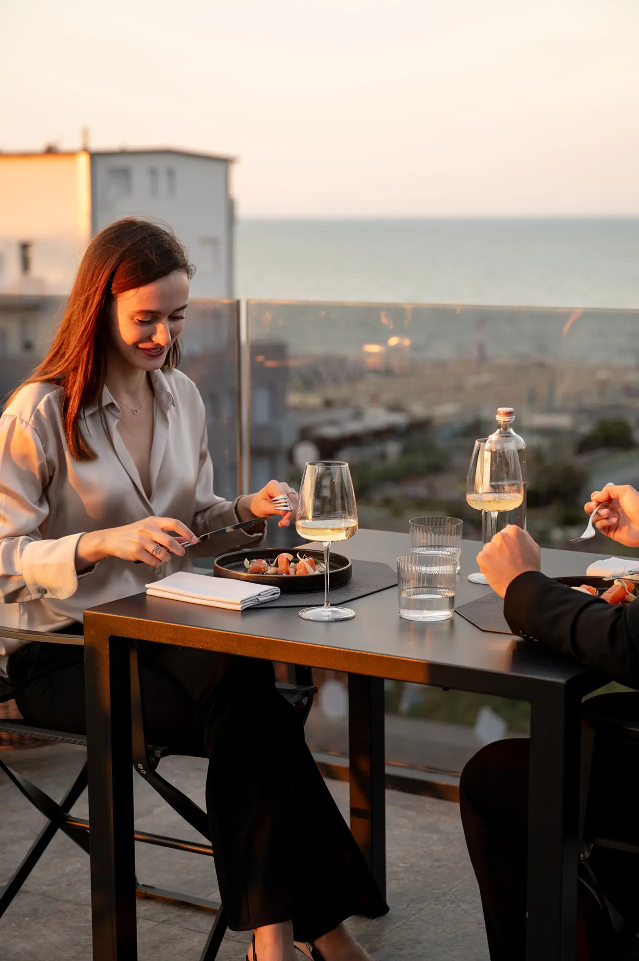 Due persone cenano all'aperto su una terrazza con vista sul mare al tramonto.