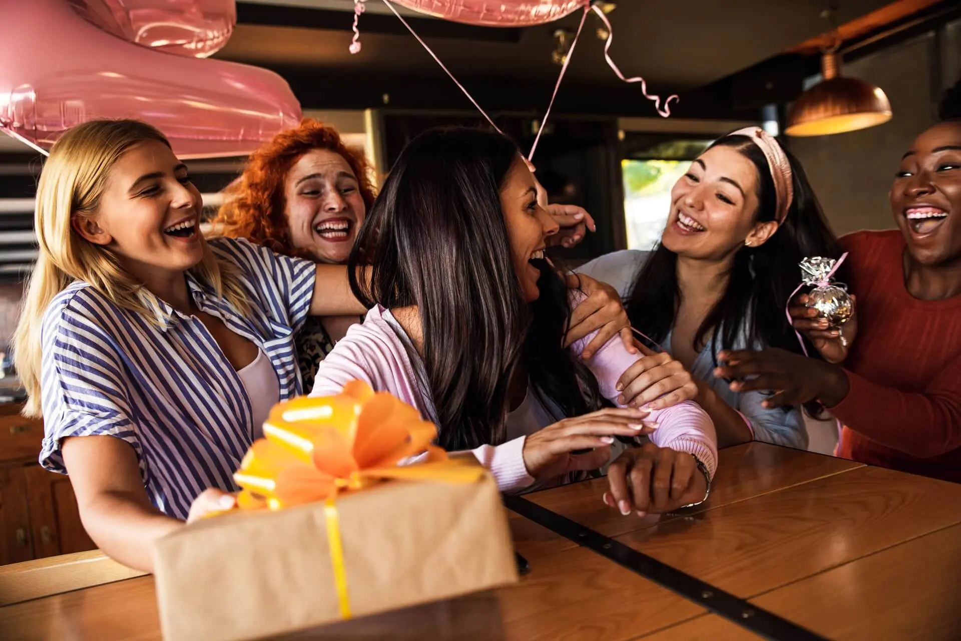 Un gruppo di amiche ride e festeggia in un locale con regali e decorazioni.