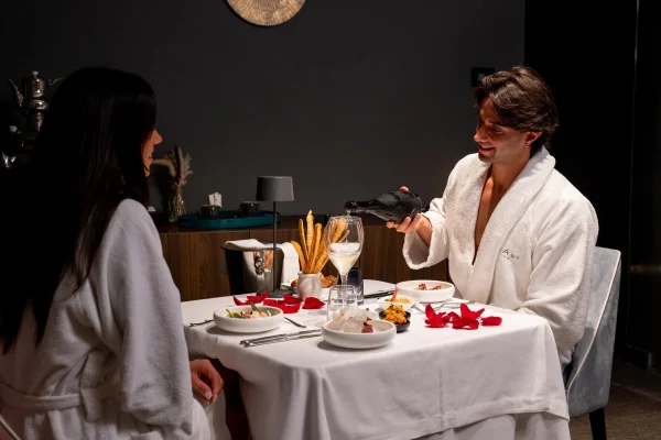 Due persone in accappatoio cenano romanticamente a un tavolo apparecchiato con petali di rosa.