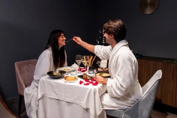 Coppia in accappatoio che cena romanticamente a un tavolo apparecchiato con petali di rosa.