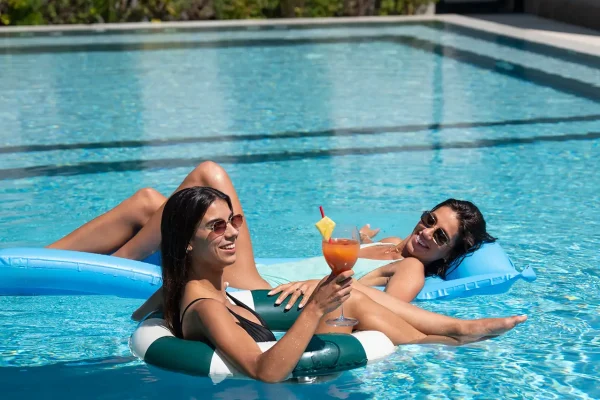 Due persone si rilassano su gonfiabili in piscina sorseggiando un cocktail sotto il sole.