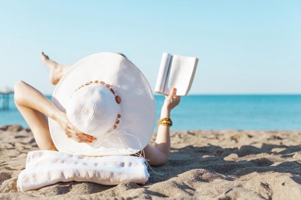 Donna con cappello bianco legge un libro sulla spiaggia al sole.