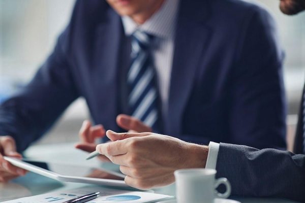 Due uomini in abiti formali discutono documenti e grafici a un tavolo con una tazza di caffè.