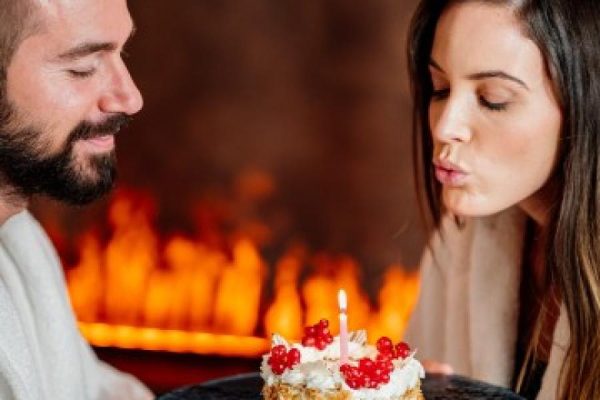 Donna che spegne una candela su una torta sorretta da un uomo, con un camino acceso sullo sfondo.
